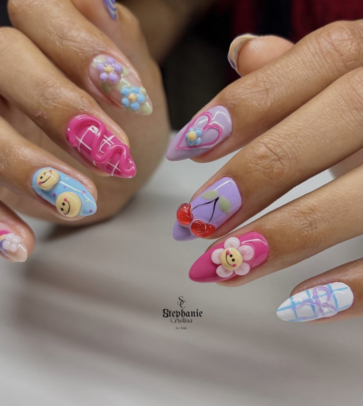 Nail art colorida com tons de rosa, lilás e azul, flores e aplicações em relevo